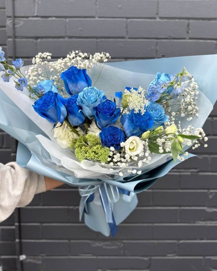 Bouquet of deep and light blue roses with greenery and white accents, wrapped in blue and white paper – elegant floral arrangement.