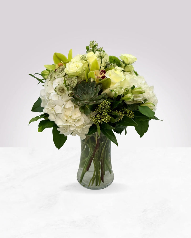 A vase arrangement of white hydrangea, roses, orchids, and green foliage.