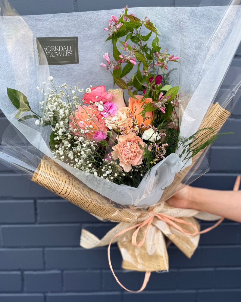  Bouquet featuring pink, peach, and orange blooms wrapped in a white and newspaper style paper.