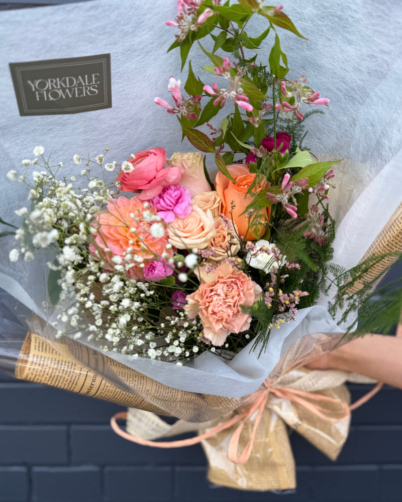  Bouquet featuring pink, peach, and orange blooms wrapped in a white and newspaper style paper.