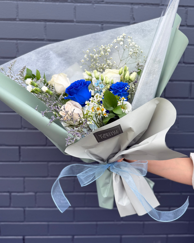 Bouquet of blue roses and white flowers, wrapped in light green paper with a ribbon.