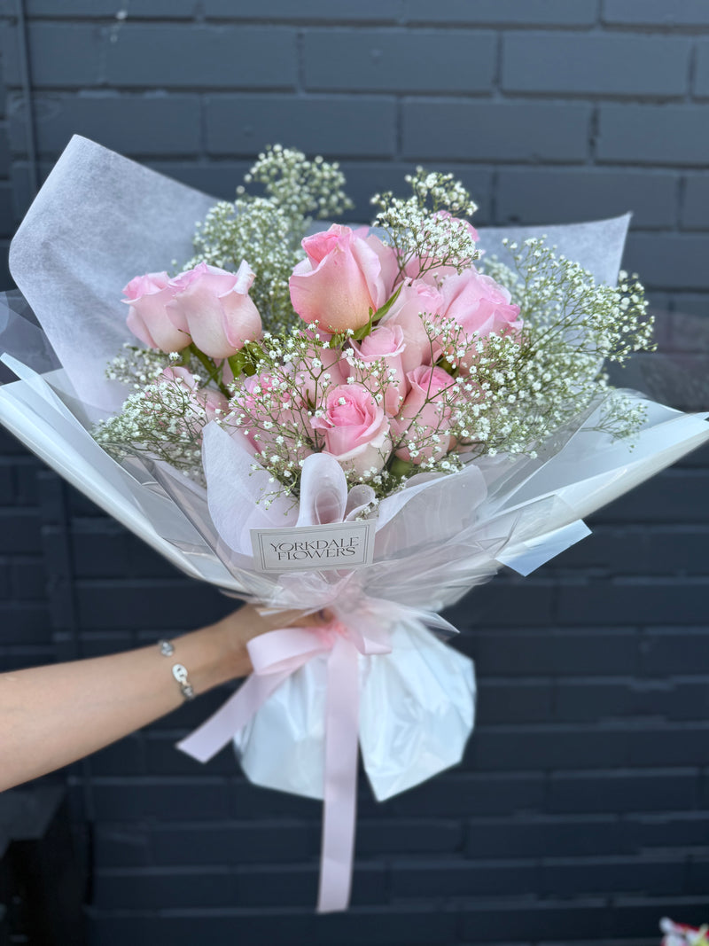 Bouquet of soft pink roses complemented by baby’s breath, wrapped in white paper with pink ribbon.