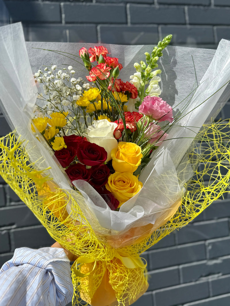 Autumn-inspired bouquet with golden yellow, deep red, and orange blooms, wrapped in white paper and yellow netting – vibrant floral arrangement.