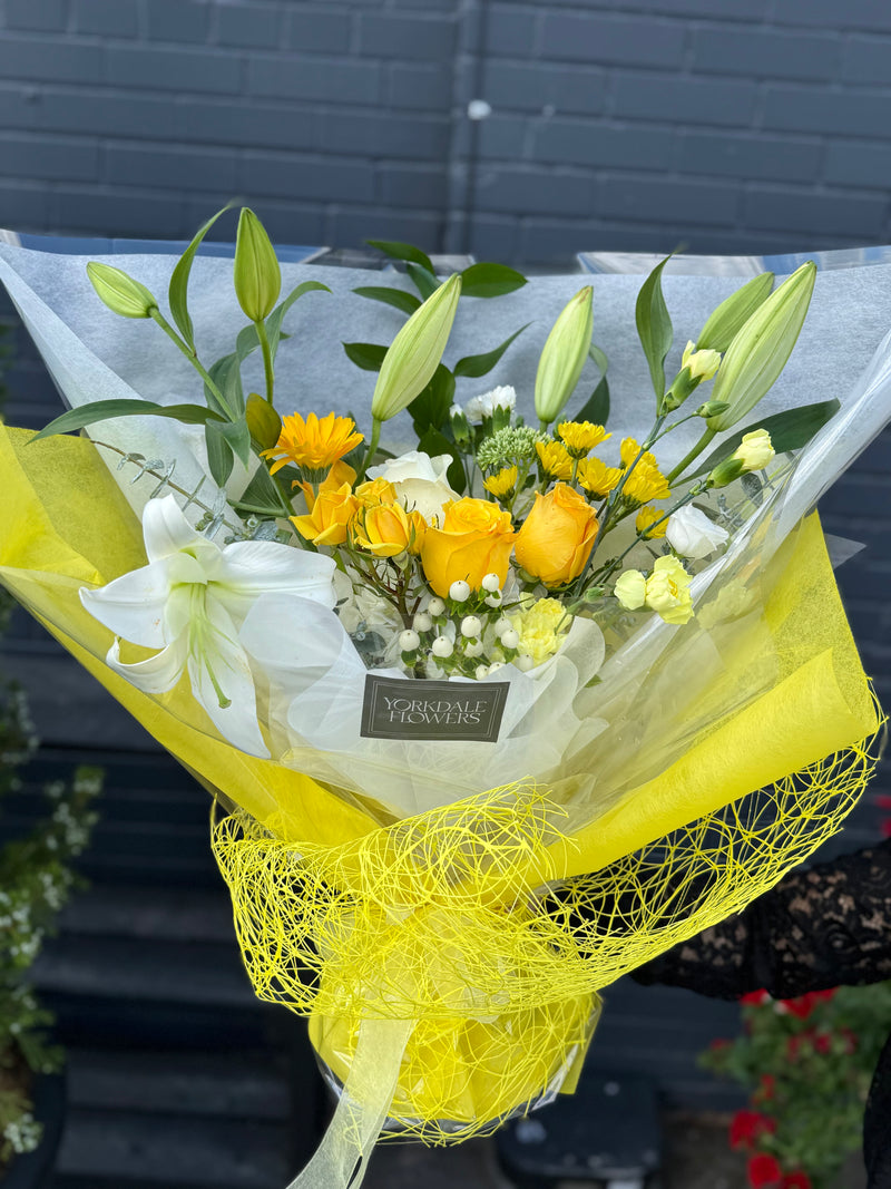 Bright yellow blooms with elegant white lilies, wrapped in white paper and yellow netting – cheerful floral bouquet.