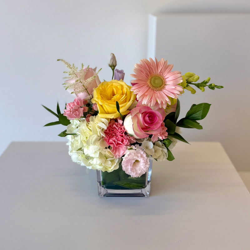 Colourful floral arrangement in a clear vase with soft pink and white blooms, accented with cheerful yellow – romantic centrepiece or floral gift.
