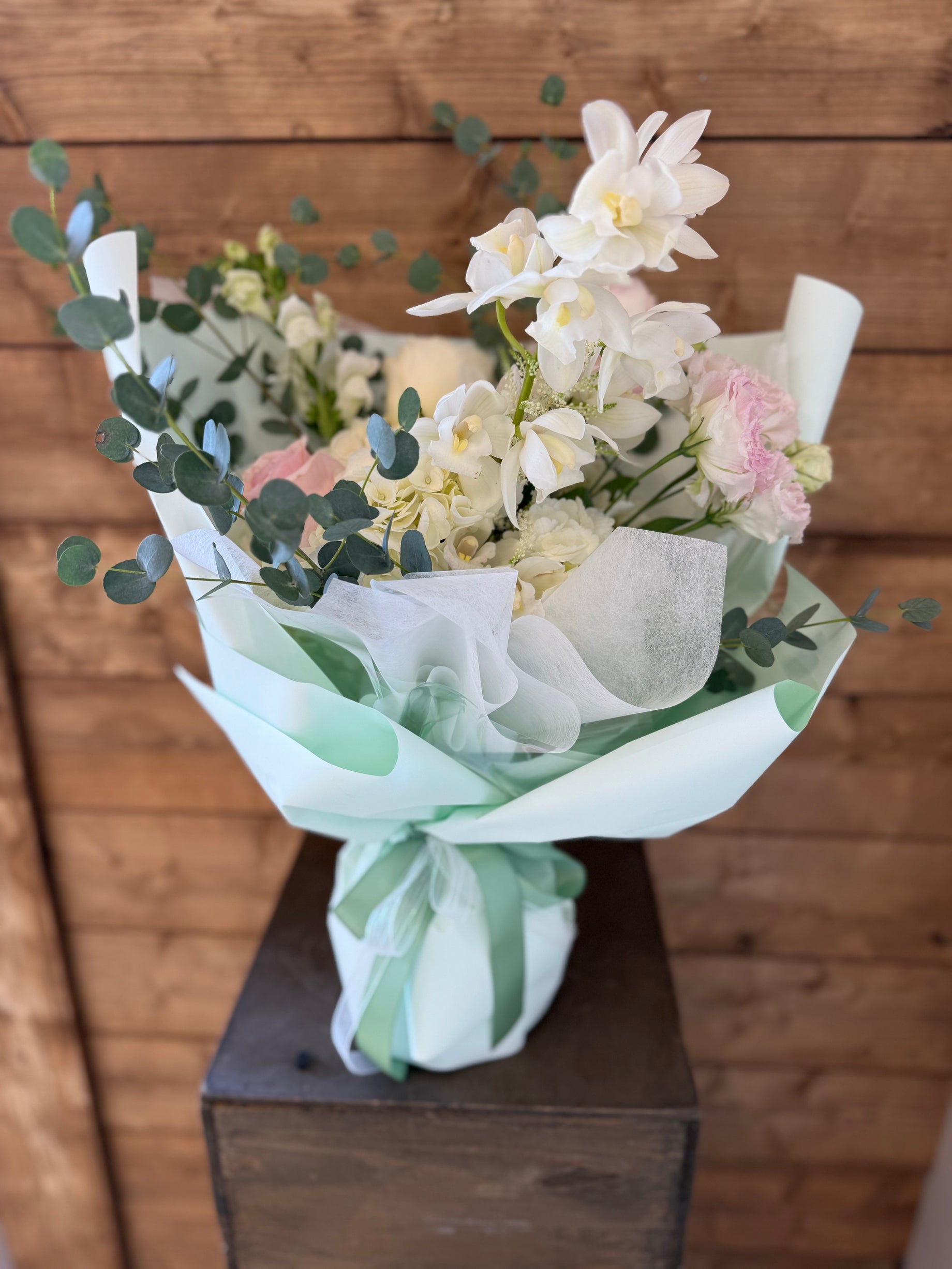 Delicate bouquet of blush pink and white orchids with mixed flowers and eucalyptus, finished in mint wrapping and tied with green ribbon – fresh floral gift arrangement.