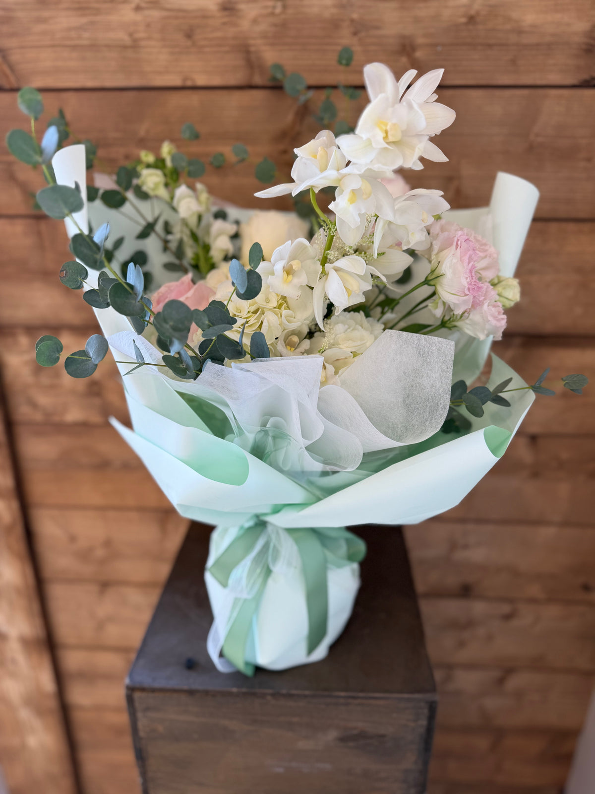Delicate bouquet of blush pink and white orchids with mixed flowers and eucalyptus, finished in mint wrapping and tied with green ribbon – fresh floral gift arrangement.