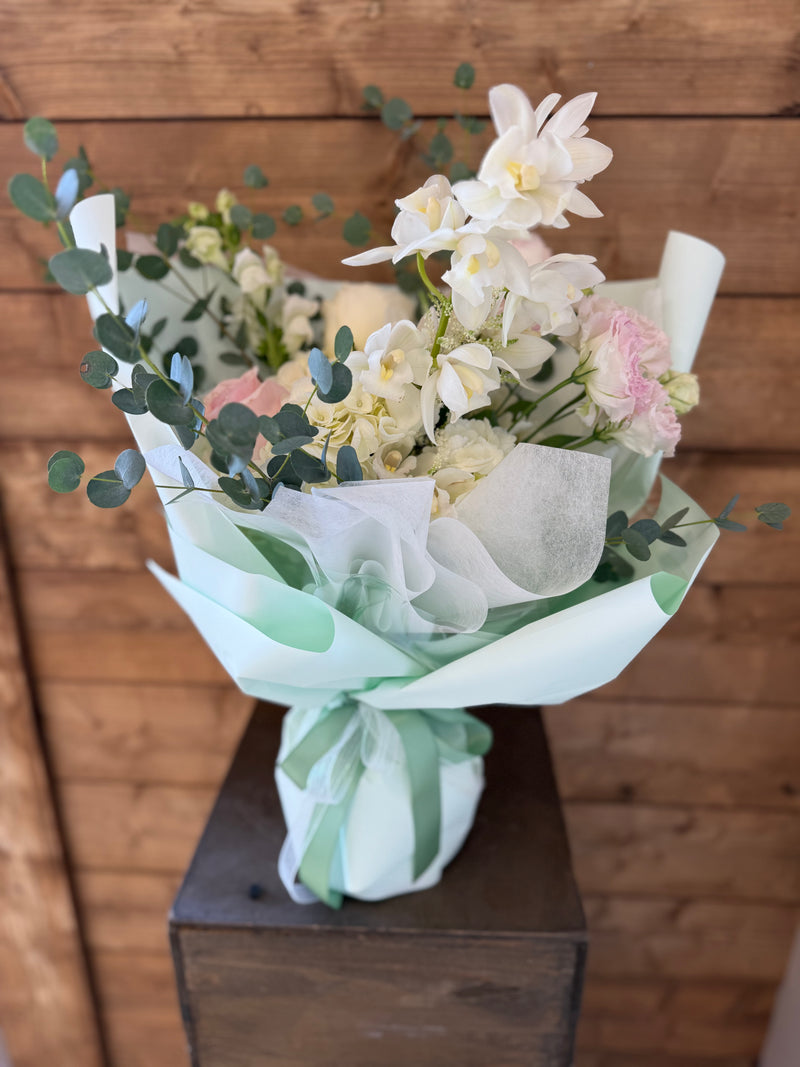 Delicate bouquet of blush pink and white orchids with mixed flowers and eucalyptus, finished in mint wrapping and tied with green ribbon – fresh floral gift arrangement.