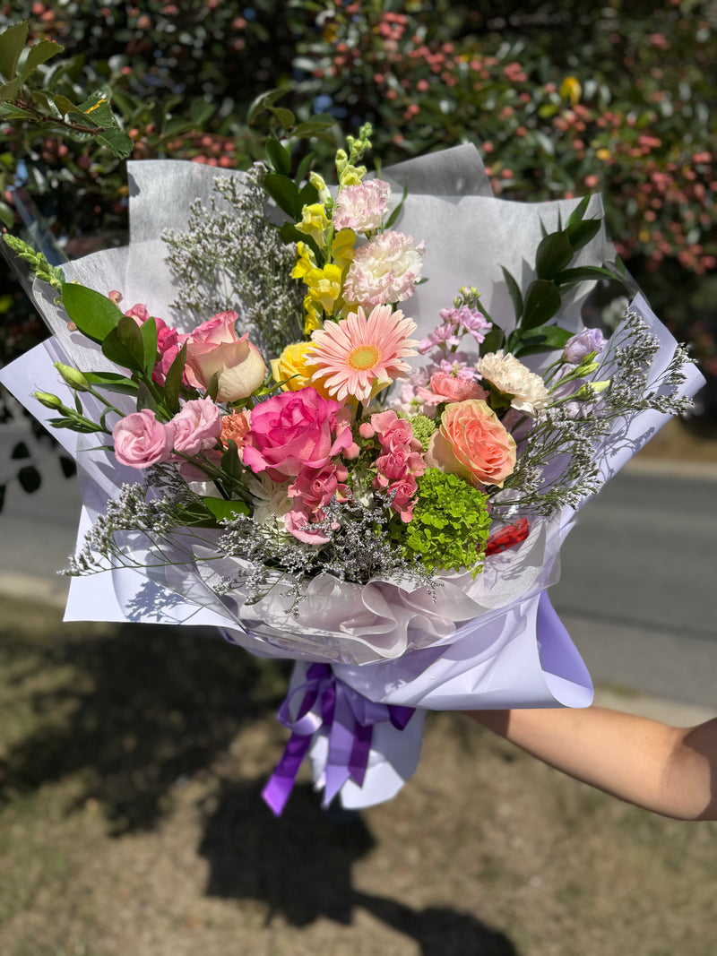 Playful bouquet with pink blooms, fresh greenery, and yellow accents, wrapped in lavender paper with purple ribbon – cheerful floral gift.
