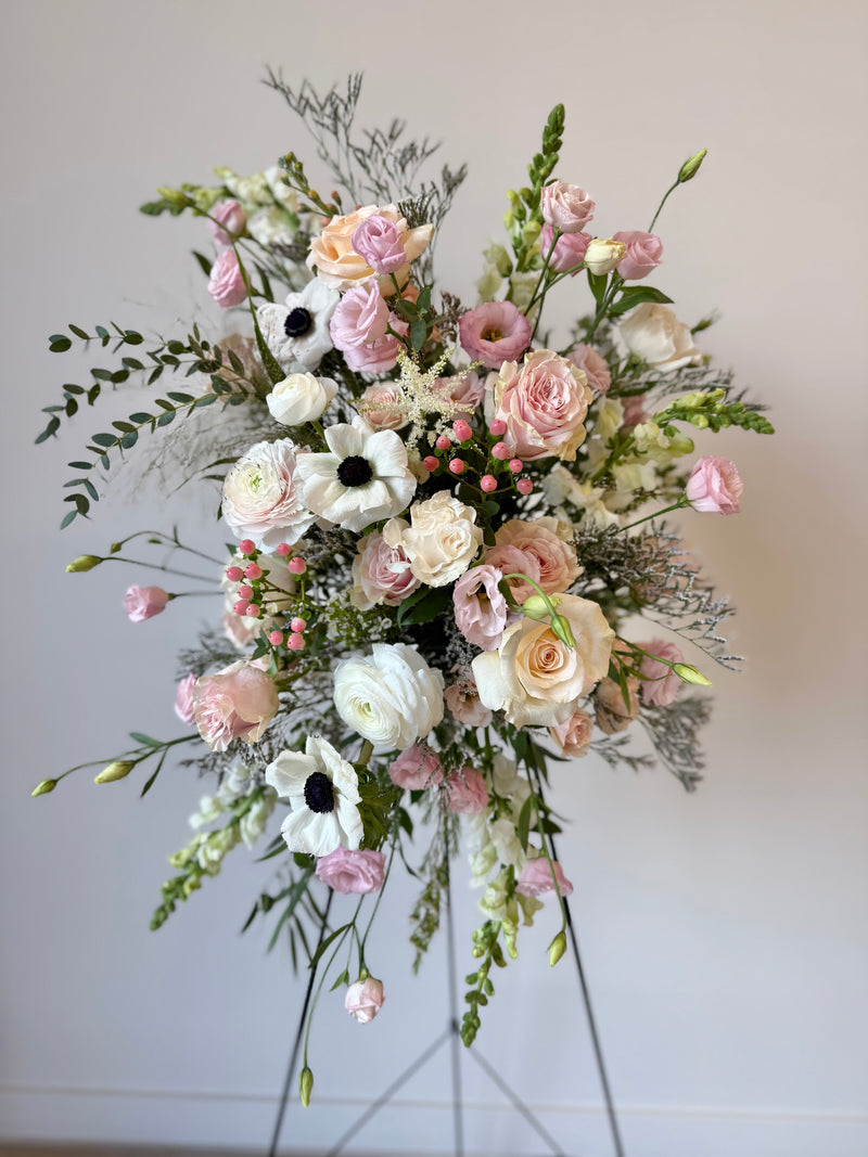 Standing funeral spray with soft pink and white blooms accented with greenery – sympathy and memorial flowers.