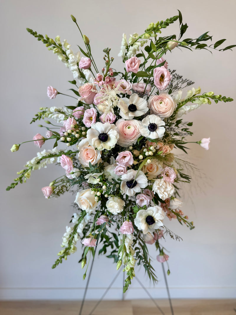 Standing funeral spray with soft pink and white blooms accented with greenery – sympathy and memorial flowers.