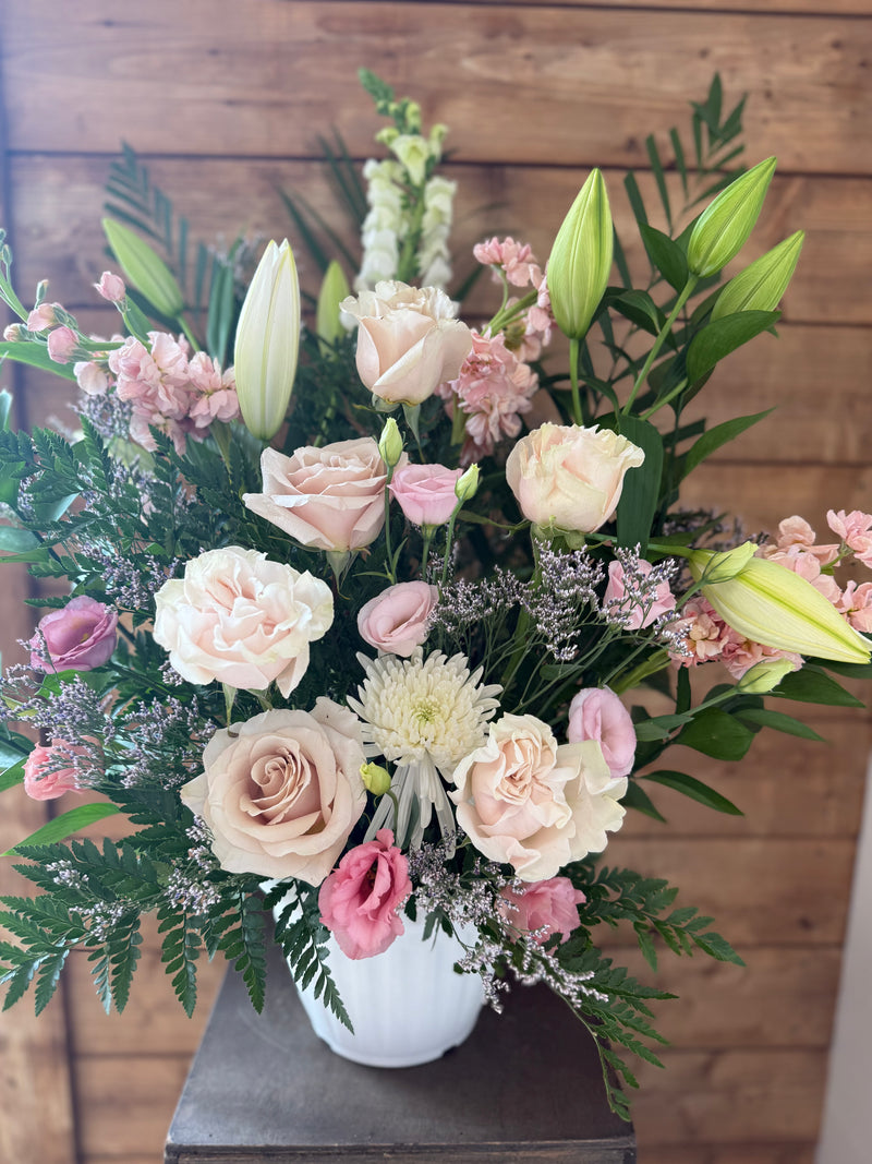 White and pale pink flowers with lilies and greenery arranged in a white vase.