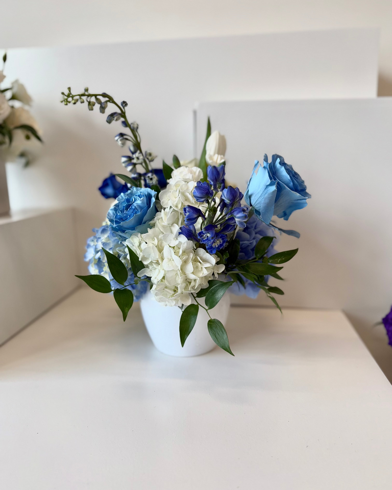 White and blue floral arrangement design with hydrangeas and roses in a white vase.