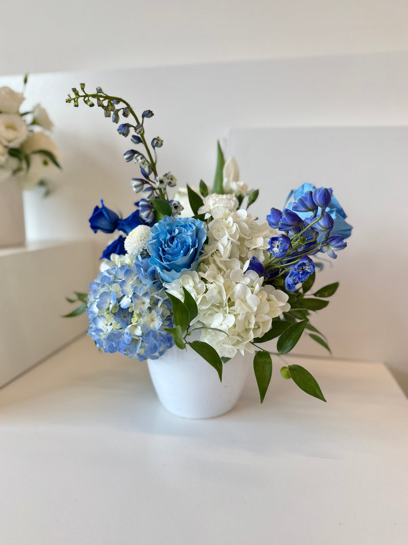 White and blue floral arrangement design with hydrangeas and roses in a white vase.