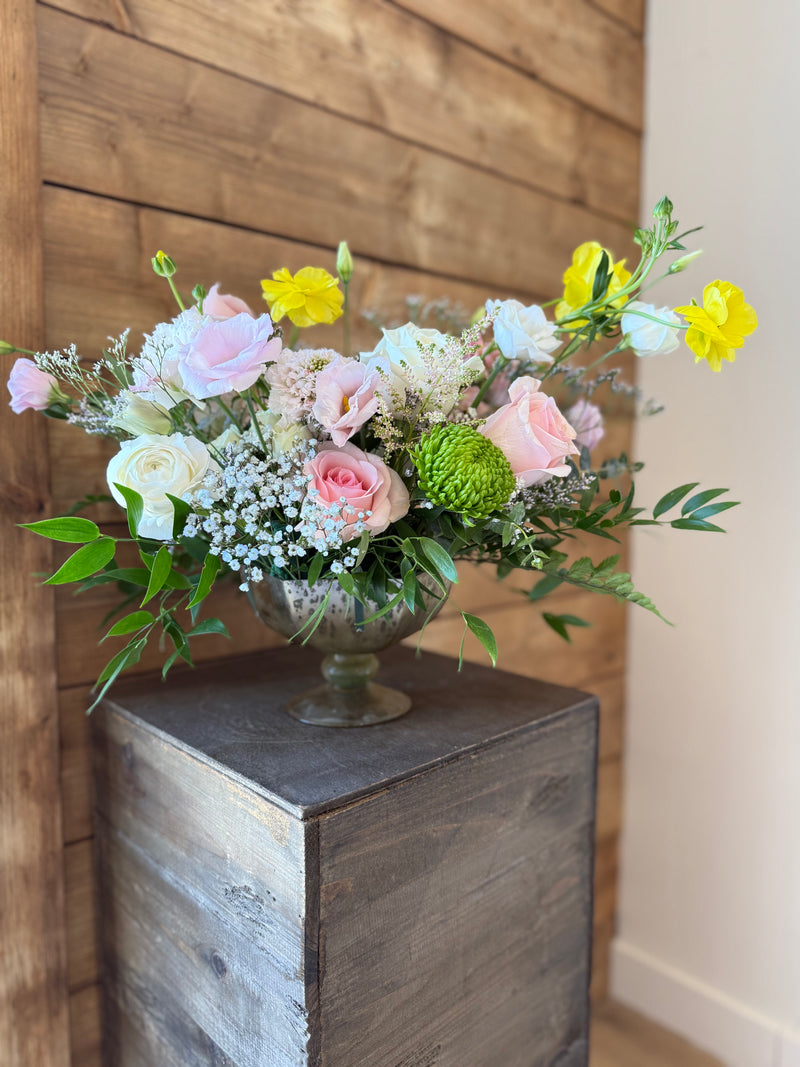 Mix of white, blush, and sunny yellow blooms with fresh greenery in a silver chalice vase.