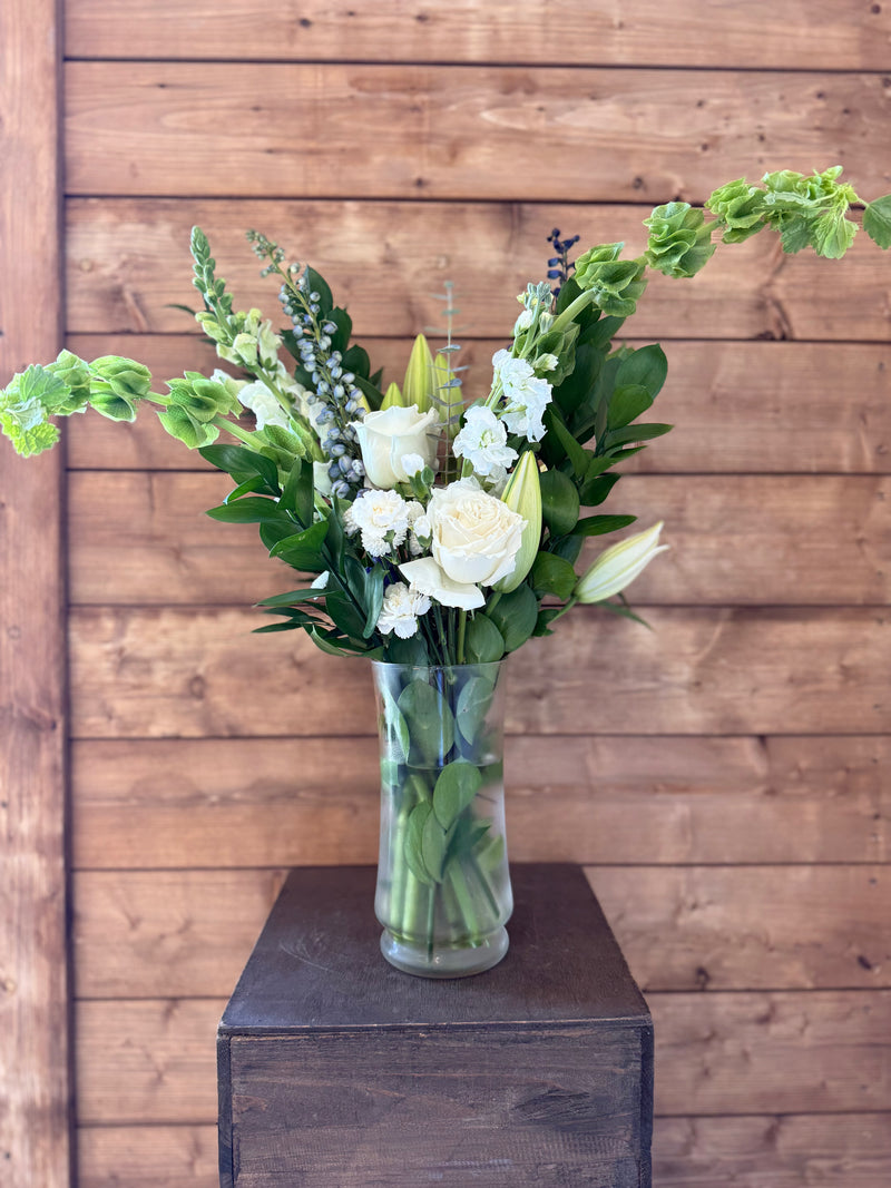 Whites and greens large brightened  flowers with blue accents, in a clear vase.