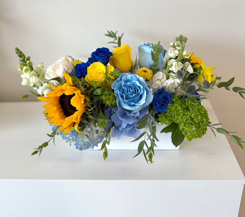 Centerpiece with a natural blend of airy blues, fresh greens, and clean whites, brightened by a cheerful sunflower in a white rectangular vase.