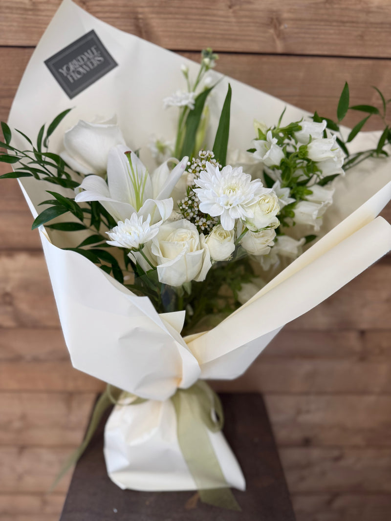 Elegant white bouquet with roses, lilies and greenery, wrapped in soft ivory paper.