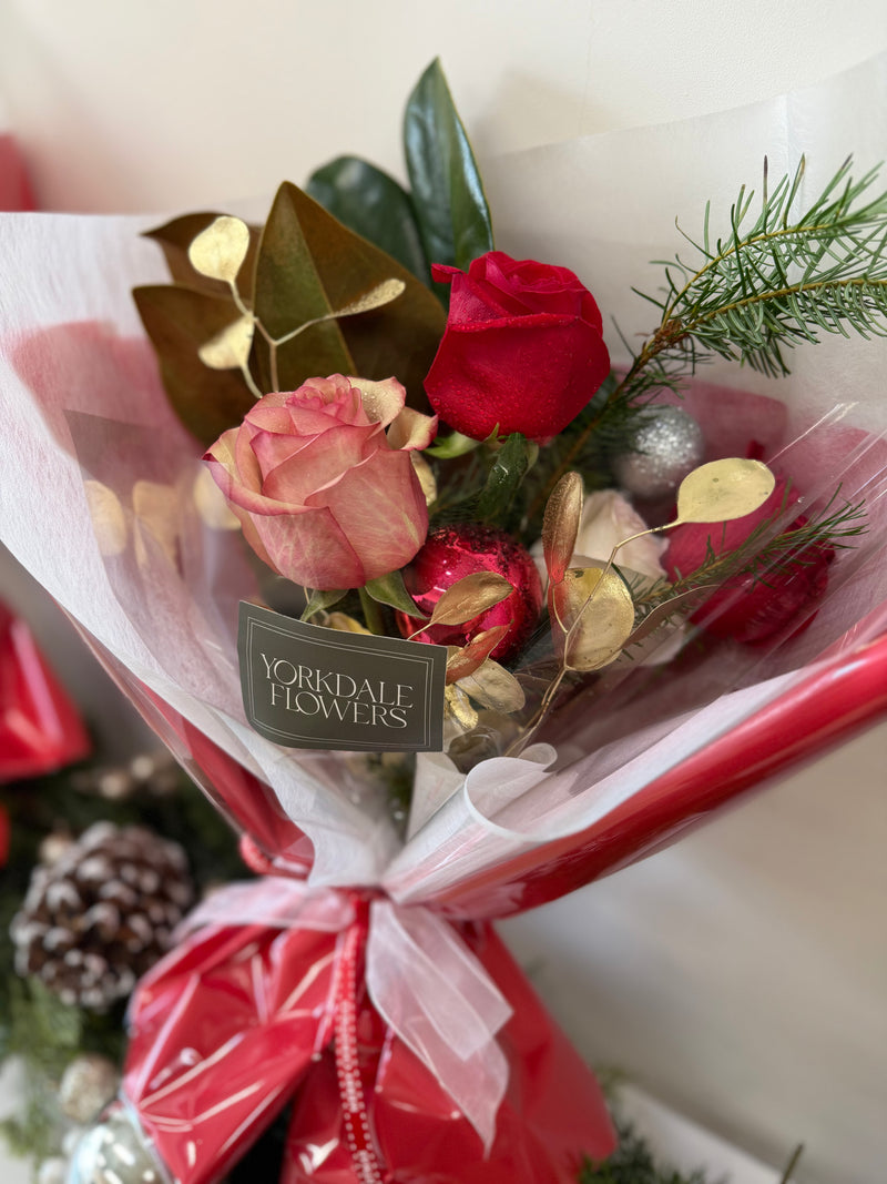 Bouquet of mix red blooms and winter foliage, finished with a delicate gold accent for a warm holiday touch.