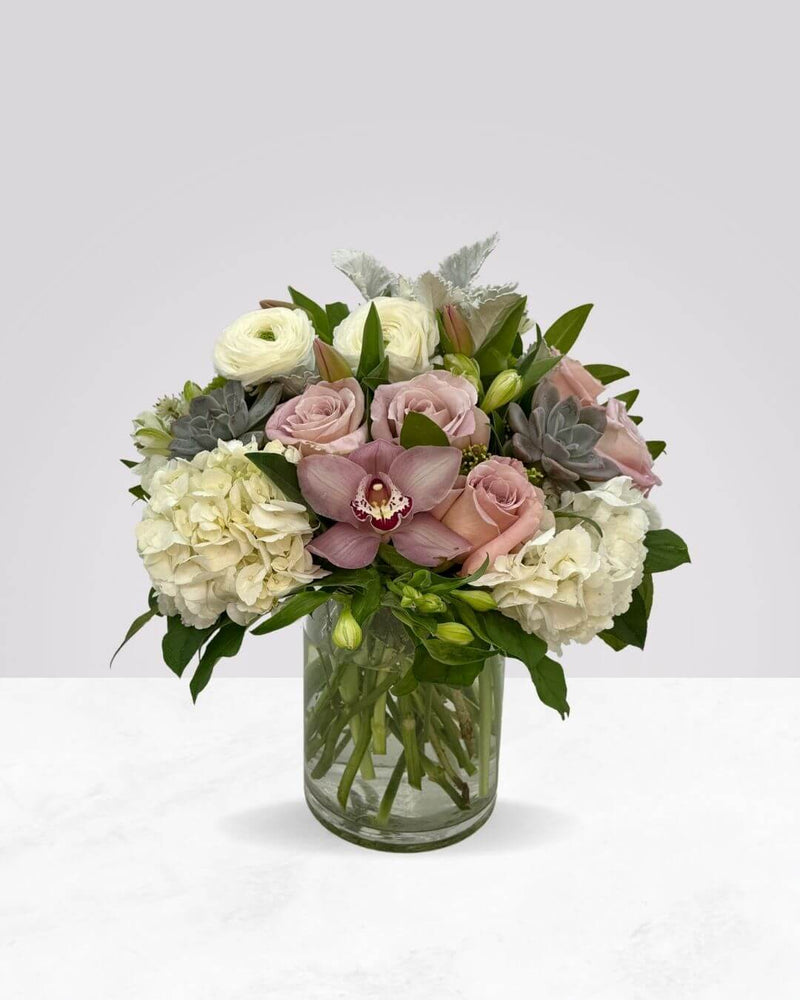 Floral arrangement featuring roses, orchids, hydrangeas, and succulents in a clear glass vase, with a mix of pink, white, and green colors.