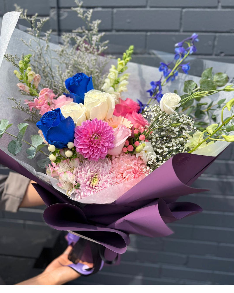 Bouquet with bright fuchsia and vivid blue blooms among soft whites and gentle pinks, wrapped with elegant purple paper.