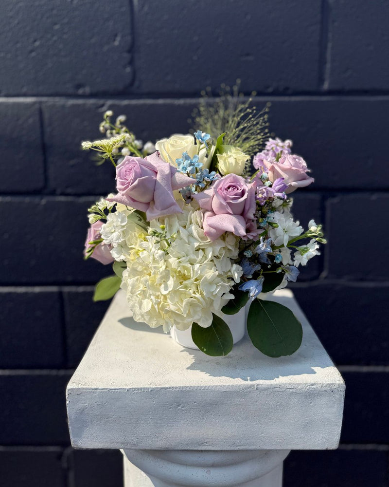 Centrepiece with touches of pale blue tucked between lavender and white blossoms in a withe cylinder vase.