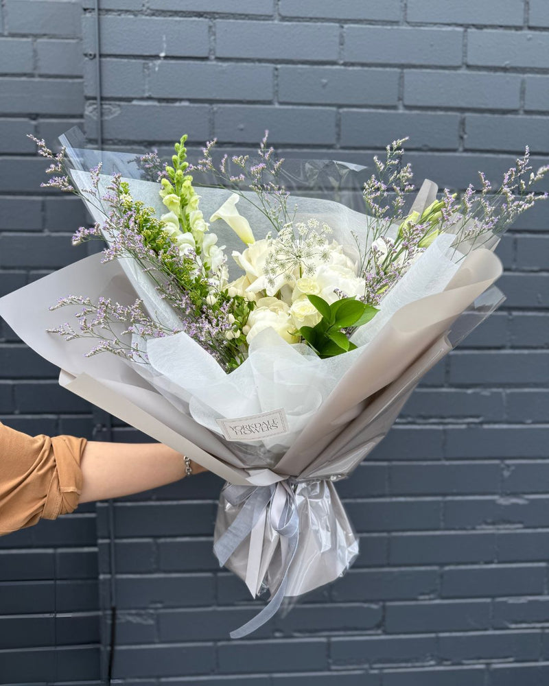 White mixed flower bouquet with greenery and lavender accents, wrapped in creamy paper – elegant floral gift arrangement.
